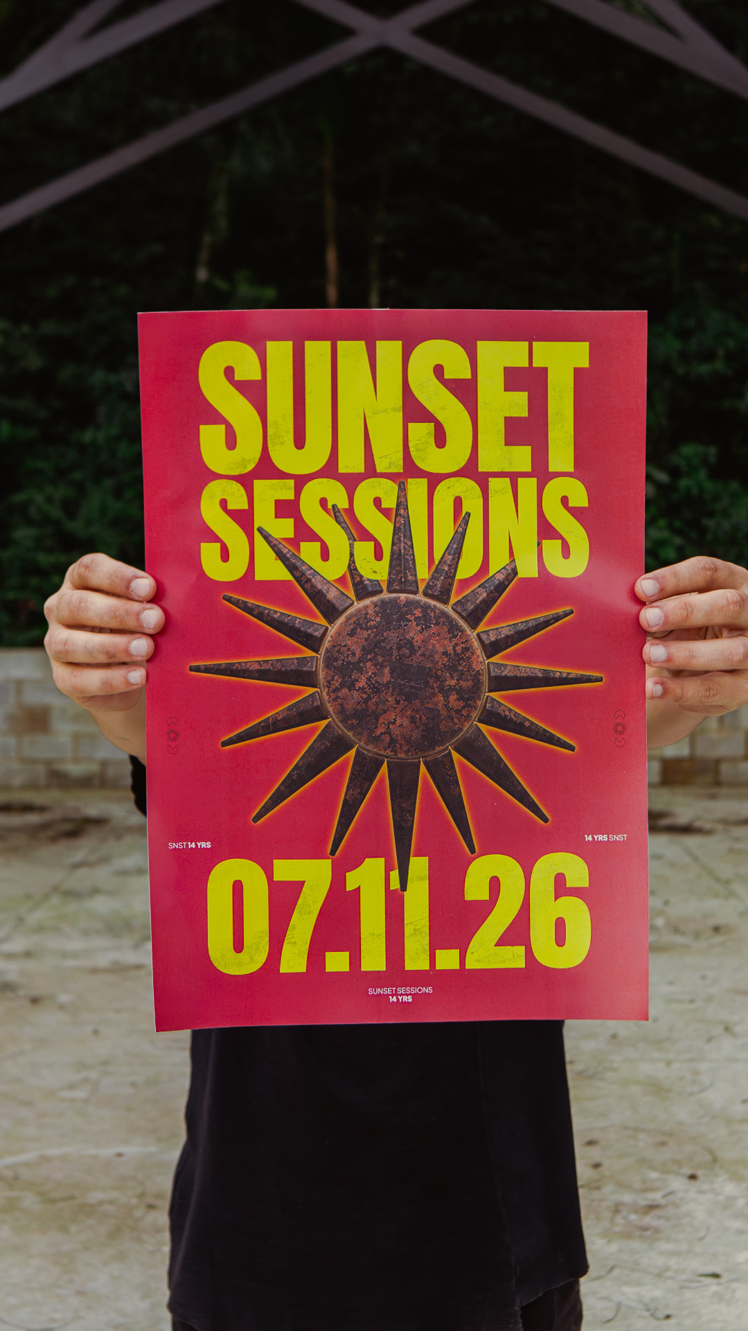 Pessoa de corpo inteiro segurando o cartaz da Sunset Sessions 14 anos